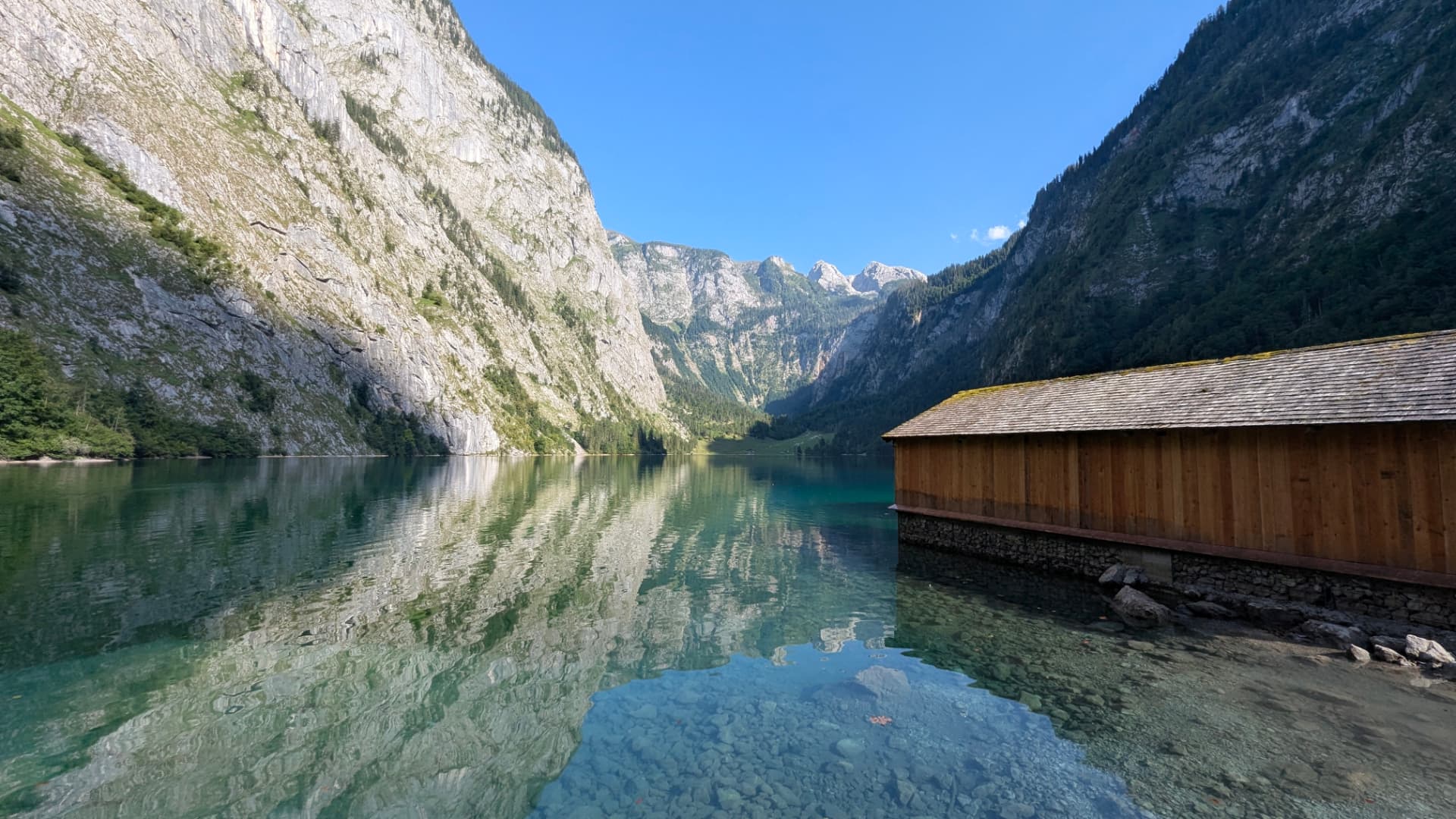Obersee lake near Königssee, with steep Alpine cliffs reflected in clear water and a wooden boathouse on the shore.