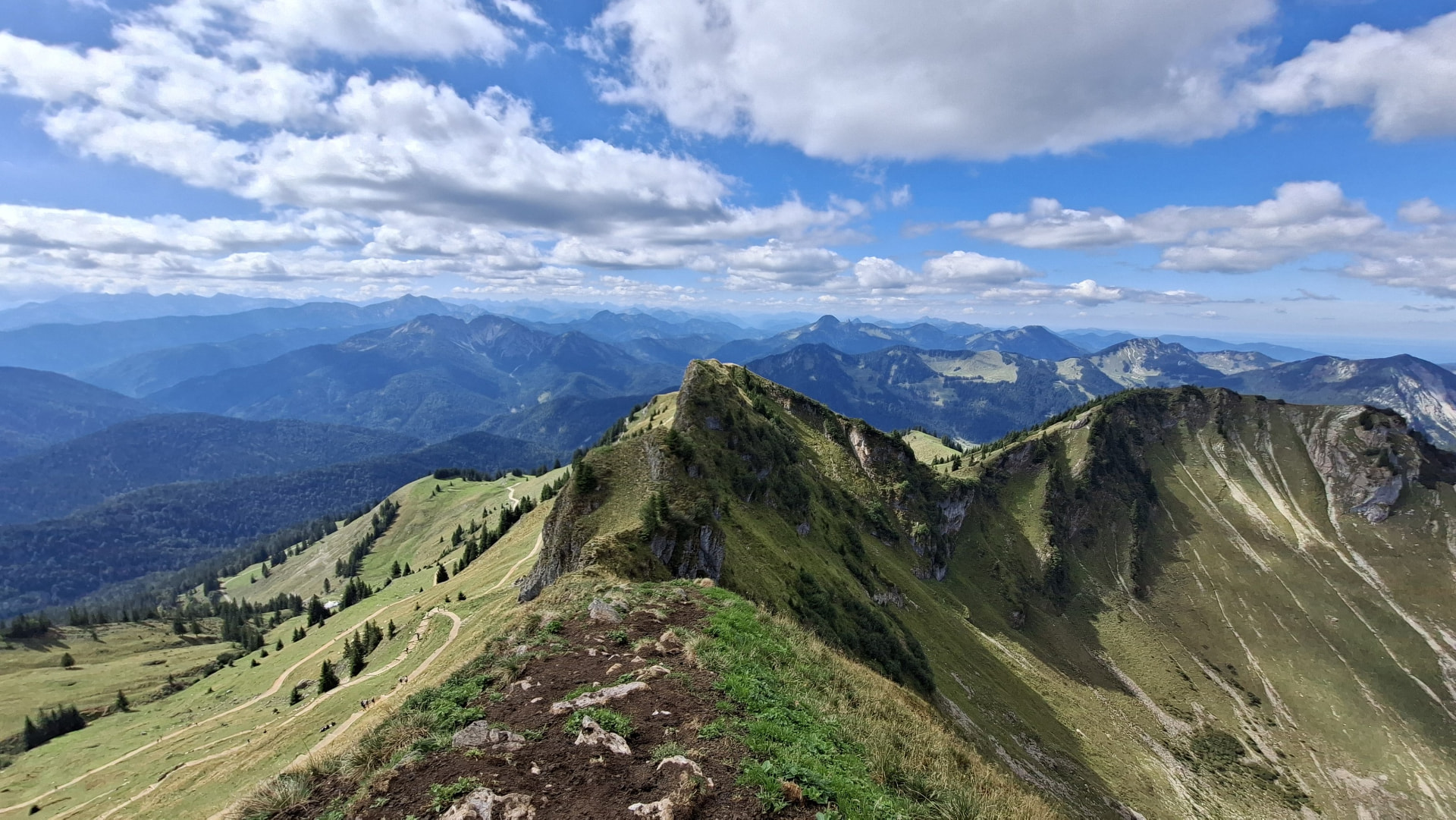 Panoramic view along the narrow Rotwand summit ridge, with hikers on the trail and layered mountain ranges in the background.